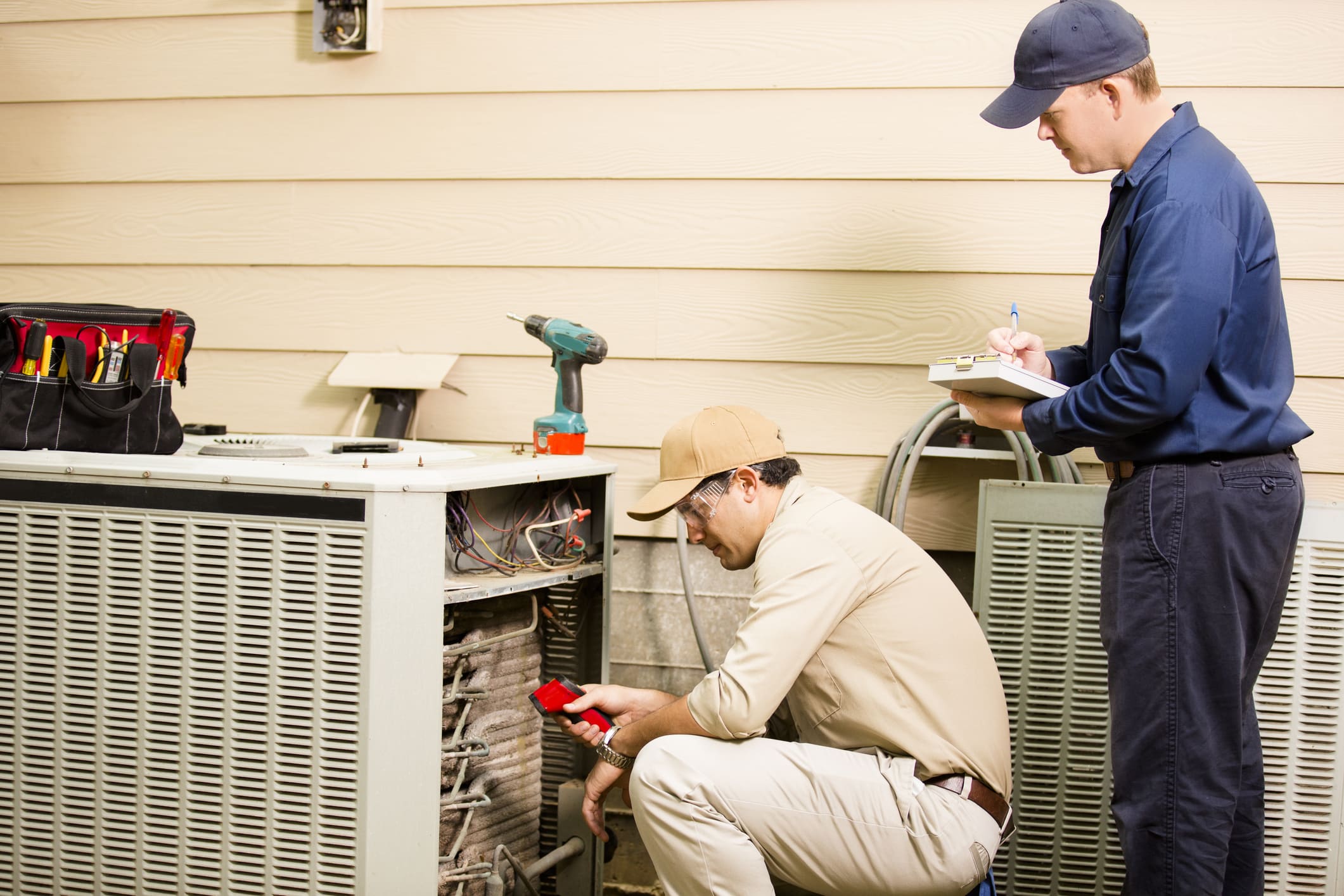 Caucasian and latin descent repairmen work on a home's air conditioner unit outdoors. They are checking the temperature and taking notes with clipboard.